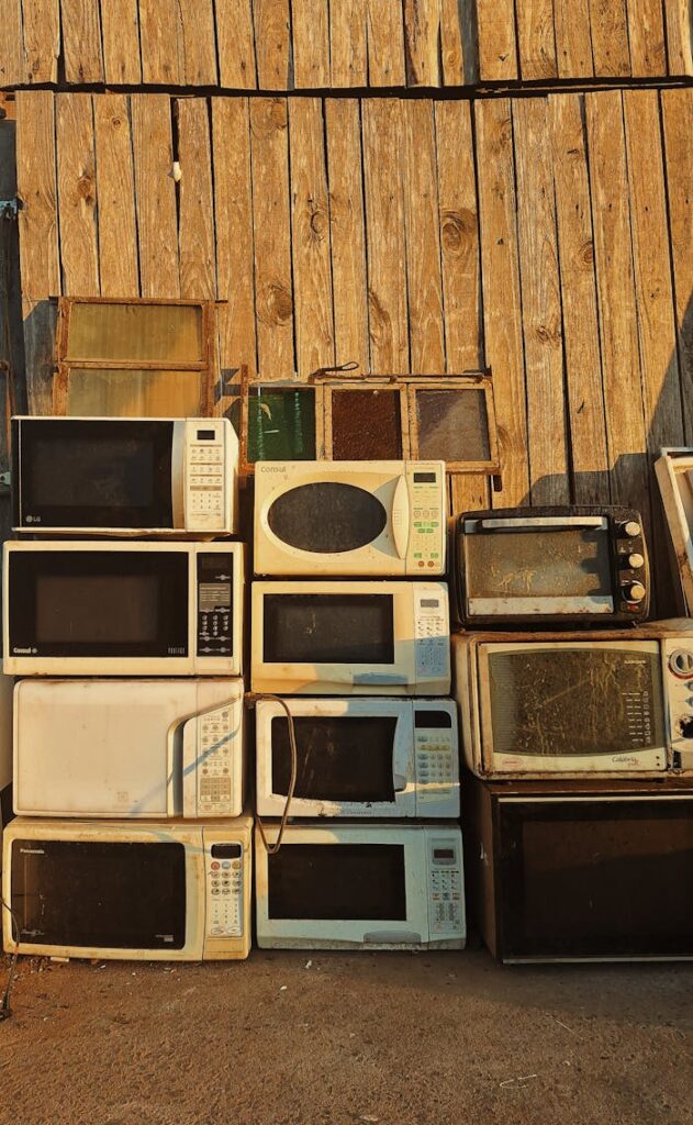 A stack of old microwaves placed outdoors against a rustic wooden wall in warm sunlight.